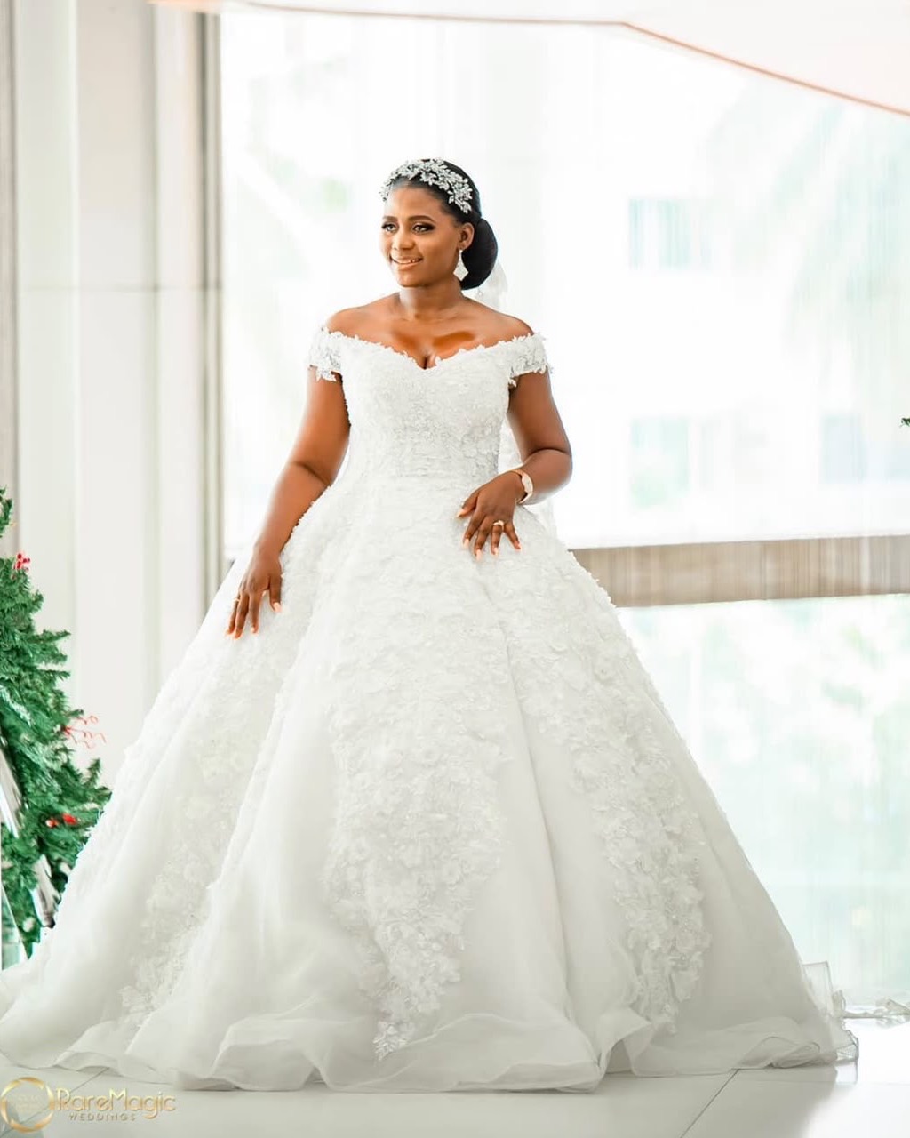 A bride in a full-skirted white lace gown and beaded headpiece, photographed in a sunlit glass-walled room