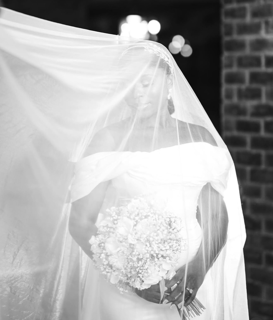 A bride beneath a flowing veil holding a bouquet, photographed in black and white